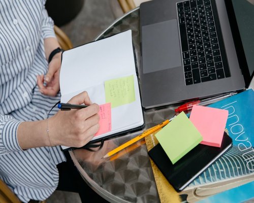 Person writing in a physical notebook with a pen, laptop closed nearby
